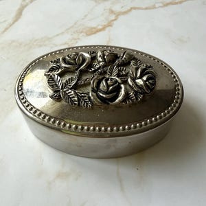 May include: Oval silver-toned jewelry box with a raised floral design on the lid. The lid features a cluster of roses and leaves. The box has a beaded border and a polished finish, suitable for storing small items.