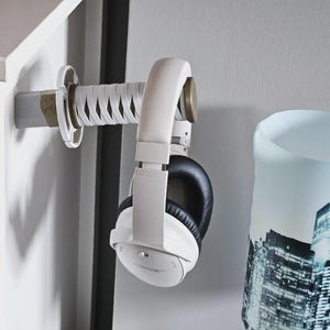 May include: A white headphone holder shaped like a katana sword is mounted on a white wall. White headphones with black earcups hang from the holder. A cylindrical lamp with a city skyline design is to the right.
