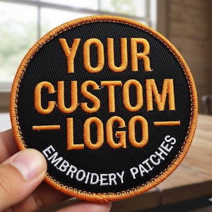 May include: Round black embroidered patch with the text "YOUR CUSTOM LOGO EMBROIDERY PATCHES" in orange thread. The patch has an orange stitched border and is held by a hand.
