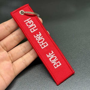 May include: A red rectangular keychain tag with the embroidered text "REMOVE BEFORE FLIGHT" in white. The tag has a metal ring for attachment. The tag is held in a hand.