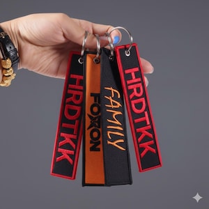 Puede incluir: Un conjunto de llaveros con texto bordado. Dos llaveros son negros con texto rojo, uno es naranja con texto negro y otro es negro con texto naranja. El texto incluye las palabras "HRDTKK", "FOXON" y "FAMILY".