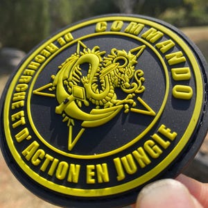 Könnte beinhalten: Ein runder, schwarzer Aufnäher mit gelbem Text und einem gelben Drachendesign. Der Text lautet "COMMANDO" und "D'ACTION EN JUNGLE". Der Aufnäher hat eine strukturierte Oberfläche.