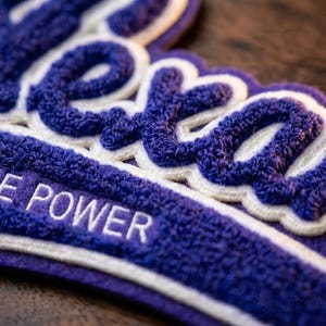 Puede incluir: Primer plano de un parche bordado morado y blanco con la palabra "Texas" en una fuente estilizada. El texto "THE POWER" está bordado en blanco debajo. El parche tiene una apariencia texturizada y en relieve.