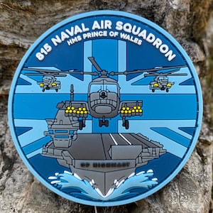 Peut inclure: Un écusson rond bleu avec le texte "815 NAVAL AIR SQUADRON HMS PRINCE OF WALES". Le motif central représente un hélicoptère, un porte-avions et le drapeau Union Jack. Les mots "OP HIGHMAST" sont en bas.