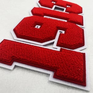 Puede incluir: Un conjunto de letras de estilo universitario en felpilla roja que forman la palabra "GSA" con un borde grueso de fieltro blanco. Las letras presentan una superficie texturizada y difusa con un diseño en relieve y un contorno blanco.