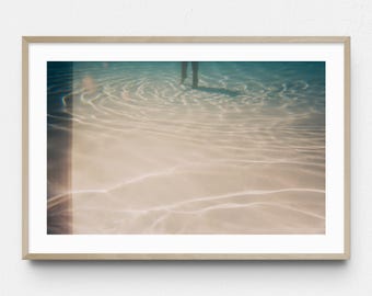 May include: A framed art print featuring a serene underwater scene. The image captures the play of light and water, with ripples and reflections creating a sense of depth. The color palette is dominated by soft blues and sandy tones, evoking a tranquil atmosphere.