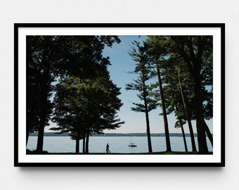 May include: A framed landscape print featuring a runner silhouetted against a lake. Tall green trees frame the scene, with a sailboat in the distance under a clear blue sky. The print is in a black frame.