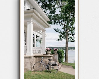 May include: A framed print featuring a white house with a porch, a bicycle, and a tree. The house has a porch with white columns and a railing. A vintage bicycle is parked on the grass. Pink flowers are in pots. A lake is visible in the background.