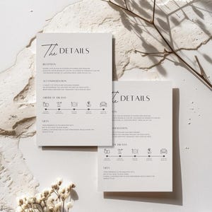 May include: Two ivory wedding detail cards with the text "The Details" in a minimalist font. The cards include information about the reception, accommodation, order of the day, and gifts. Delicate white flowers and branches are arranged around the cards.