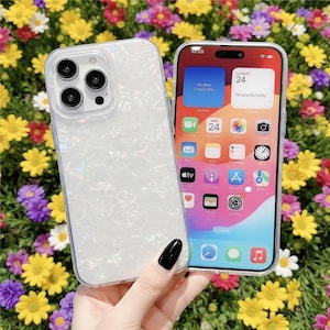 May include: A clear phone case with a pearlescent, iridescent design. The phone case is next to a smartphone with a colorful display showing the date as Sunday, the 24th. The background is a bed of colorful flowers.