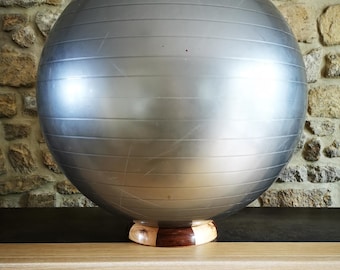 May include: A large, spherical, silver exercise ball featuring horizontal embossed lines across its surface. The ball is supported by a circular wooden base constructed from alternating light and dark wood segments, providing stability for the sphere.