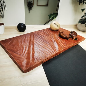 Handmade buffalo leather gym mat displayed with vintage sports accessories on a wooden floor. The full-grain leather surface, warm brown patina, and artisanal stitching make this decorative tatami perfect for industrial loft interiors or elegant home gym spaces. Crafted in France by Ben & Flo with premium materials.