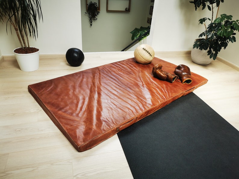 Handmade buffalo leather gym mat displayed with vintage sports accessories on a wooden floor. The full-grain leather surface, warm brown patina, and artisanal stitching make this decorative tatami perfect for industrial loft interiors or elegant home gym spaces. Crafted in France by Ben & Flo with premium materials.