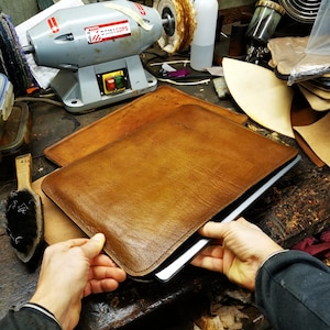 Puede incluir: Una funda para portátil de cuero marrón que se coloca sobre un portátil. La funda la sujetan las manos de una persona. La funda está sobre una mesa de madera con herramientas y otros artículos de cuero.