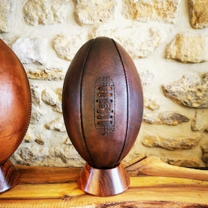 Puede incluir: Dos balones de rugby de cuero marrón sobre soportes de madera. Los balones se muestran sobre una superficie de madera con una pared de piedra en el fondo.