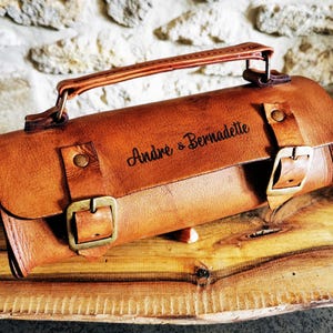 Peut inclure: Un sac enroulable en cuir marron avec une poignée et deux boucles. Le sac est personnalisé avec le texte "Andre & Bernadette".