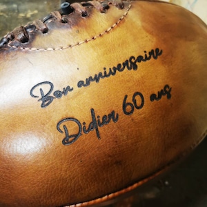 Peut inclure: Un ballon de rugby en cuir brun avec des coutures noires et le texte "Bon anniversaire Didier 60 ans" gravé sur le côté.