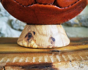 May include: A vintage-style brown leather ball with visible stitched seams resting on a circular, light-colored wooden base featuring a natural wood grain pattern and small dark knots, showcasing a smooth, polished finish on the stand.