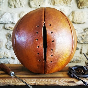 Op de afbeelding: Een bruine leren basketbal in vintage-stijl met een centrale verticale opening en voorgeponste vetergaten. De bal heeft een gladde, gepolijste textuur en wordt vergezeld door een metalen priem en een zwart leren veterkoord.