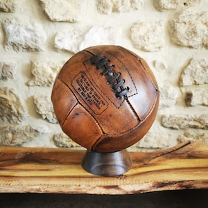May include: A vintage brown leather soccer ball with black stitching and a label that reads "HAND MADE SINCE 1902 - DESIGNED IN FRANCE - VEGETABLE TAN LEATHER - BY MEDEA & FILS"