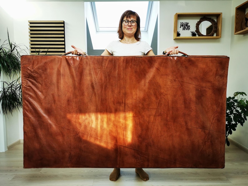Full front view of a person holding the handmade buffalo leather mat vertically to show its real size. The large 180x100cm format, elegant patina, and soft foam interior make it suitable for yoga, relaxation, or stylish home decor with a vintage touch.