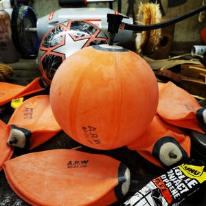 Könnte beinhalten: Ein orangefarbener aufblasbarer Ball wird mit einem schwarzen Schlauch aufgepumpt. Mehrere entleerte orangefarbene Blasen mit dem Text "A.R.W. 60-65 GM" sind verstreut. Ein Fußball und Werkzeuge befinden sich im Hintergrund.