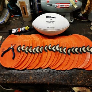 Può includere: Primo piano di una pila di 18 cuscinetti da allenamento per il football americano arancioni e neri. I cuscinetti sono disposti a semicerchio su una superficie di legno. Un paio di pinze è visibile al centro della pila.