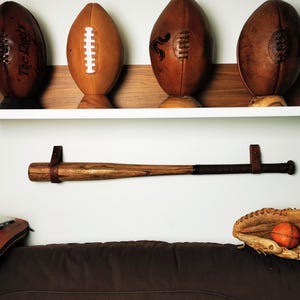 May include: A display featuring four brown leather footballs on a wooden shelf, a baseball bat with a leather strap, and a baseball glove with a ball. The items are arranged against a white wall.