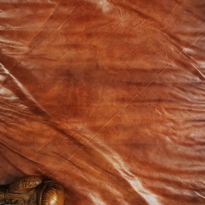 Detail shot of the buffalo leather patina with visible engraving mark and fine stitches. The soft mineral-tanned surface reflects warm brown and caramel tones, showcasing authentic handmade quality and premium craftsmanship for collectors and interior designers.