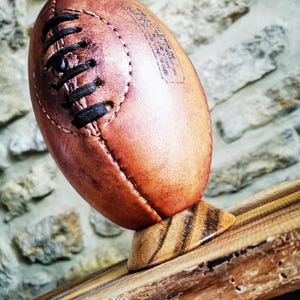 Peut inclure: Un ballon de rugby en cuir brun avec des lacets noirs posé sur un support en bois. Le ballon porte du texte blanc.