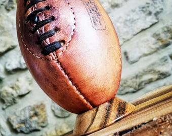 May include: A brown leather rugby ball with black laces sits on a wooden stand. The ball has white text on it.