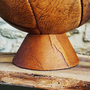 Peut inclure: Un bol sphérique en bois avec une finition texturée marron semblable à du cuir. Le bol repose sur une base conique, également marron. L'ensemble est présenté sur une surface en bois rustique.