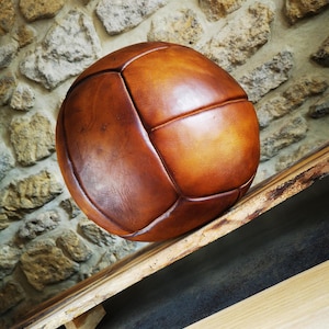 May include: A vintage-style brown leather football featuring a multi-panel construction with visible stitched seams. The ball has a smooth, polished surface with natural colour variations ranging from light tan to deep mahogany brown tones.