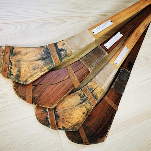 May include: Four wooden hurleys, traditional Irish sports sticks, with leather wrappings and metal nameplates. The hurleys are arranged in a fan shape, with the tips pointing towards the top of the image.