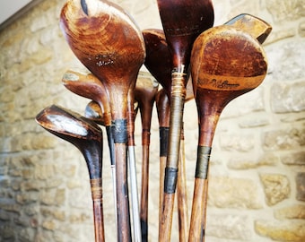 May include: A collection of vintage golf clubs with wooden heads and shafts, featuring dark brown wood grain, worn finishes, and dark tape wrappings on the handles, all standing upright inside a dark brown leather golf bag container.
