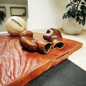 Lifestyle scene showing the vintage buffalo leather mat with old sports balls and gear beside it. The setup evokes nostalgic gym aesthetics, perfect for decorating studios, offices, or sports-themed interiors. Each Ben & Flo piece is unique and handcrafted in France.