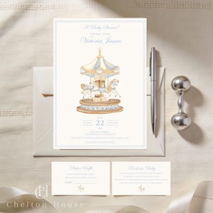 Op de afbeelding: Een uitnodiging voor een babyshower met een carrousel-illustratie in zachte blauwe en gouden tinten. De uitnodiging bevat de tekst "A Baby Shower!" en "Victoria James". Een zilveren pen en rammelaar liggen naast de uitnodiging.