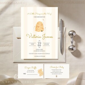 May include: Baby shower invitation with a bee theme. The invitation features the text "A Little Honey is on the Way!" and "Come Shower Mom-to-Bee". The invitation includes the name "Victoria James", date, time, and location. A silver pen and rattle are also present.