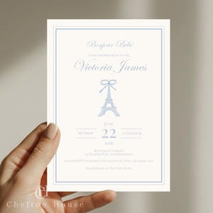 May include: A light cream-colored invitation with a blue border and text. The invitation reads "Bonjour Bébé" and "Victoria James" with an Eiffel Tower illustration. The date is June 22, 2020, at 2 o'clock. The invitation is for a baby shower.