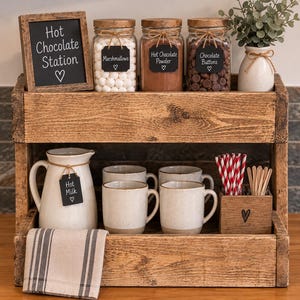 May include: A two-tiered wooden stand holding a hot chocolate station with three glass jars labelled Marshmallows, Hot Chocolate Powder, and Chocolate Buttons, a white pitcher labelled Hot Milk, four speckled mugs, a striped towel, and a sign.