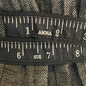May include: Close-up of a dark fabric with a herringbone pattern. Two black ribbons with white markings and text are visible. The top ribbon reads "ANNA" and the bottom ribbon reads "SUI". The ribbons have inch markings.
