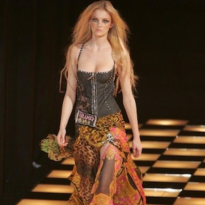 May include: A model wearing a black corset top with silver embellishments and a patterned skirt. The skirt features a leopard print and a floral design with pink and gold accents. The outfit is accessorized with a skull detail.