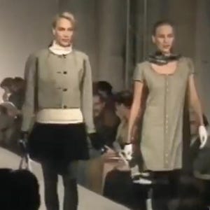 May include: Two models on a runway showcase a fashion collection. The first model wears a light green jacket over a white top and a black skirt. The second model wears a light green dress with a scarf.