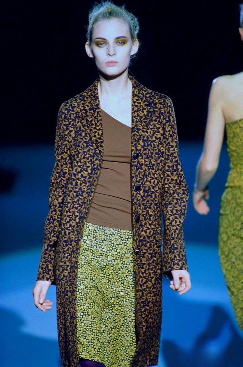 May include: A long-sleeved coat featuring a dark blue and mustard yellow floral pattern, worn over a solid brown one-shoulder top and a yellow skirt with a geometric pattern. The coat has a notched lapel and a single-breasted button closure.