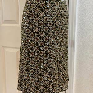 May include: A knee-length skirt featuring a black base with a repeating geometric pattern in yellow and dark red. The fabric is embellished with small, scattered silver sequins throughout the design, creating a subtle reflective texture across the surface.