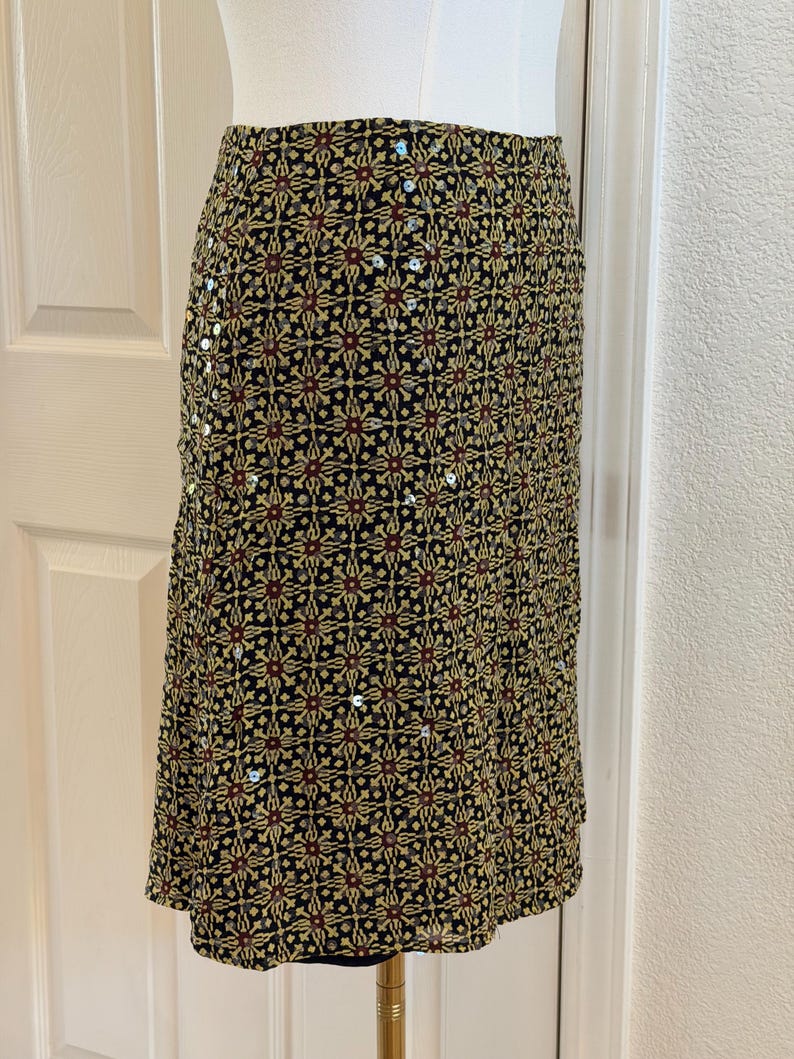 May include: A knee-length skirt featuring a black base with a repeating geometric pattern in yellow and dark red. The fabric is embellished with small, scattered silver sequins throughout the design, creating a subtle reflective texture across the surface.