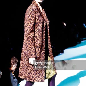May include: A knee-length coat featuring a dark brown and black star-patterned print, worn over a solid black garment with a visible gold-toned textured hem. The ensemble is paired with opaque dark purple tights and dark footwear.