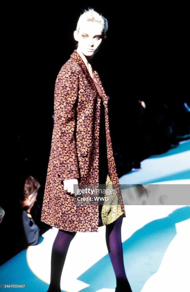 May include: A knee-length coat featuring a dark brown and black star-patterned print, worn over a solid black garment with a visible gold-toned textured hem. The ensemble is paired with opaque dark purple tights and dark footwear.