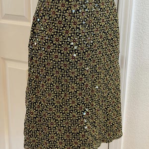 May include: A knee-length skirt featuring a black base with a repeating geometric pattern in yellow and dark red. The fabric is embellished with small, scattered silver sequins that catch the light, creating a subtle shimmer across the surface.