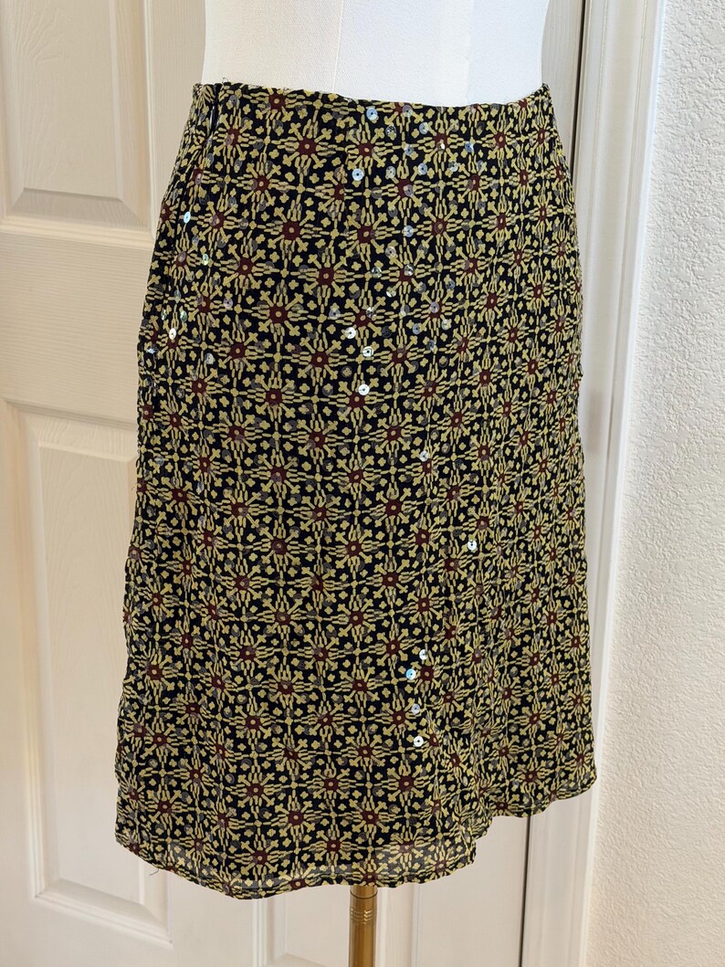 May include: A knee-length skirt featuring a black base with a repeating geometric pattern in yellow and dark red. The fabric is embellished with small, scattered silver sequins that catch the light, creating a subtle shimmer across the surface.
